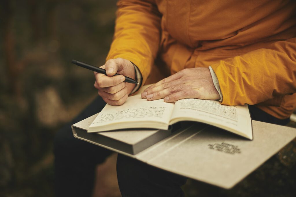 sitzende Person mit Buch auf den Knien und Stift in der Hand